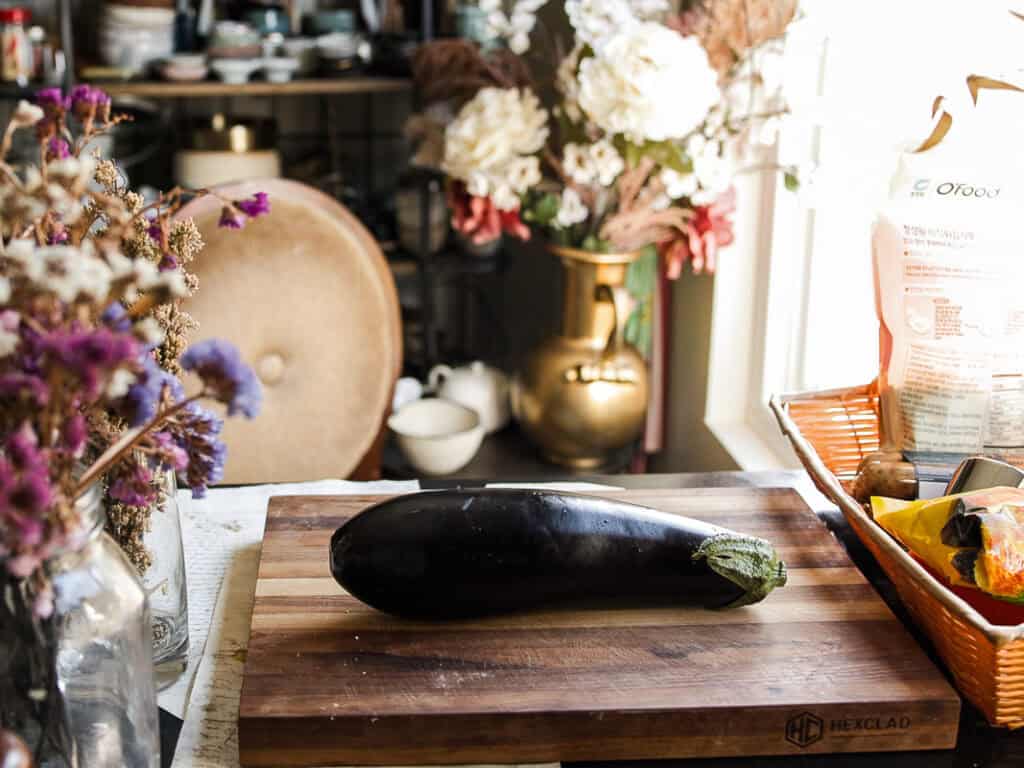 An eggplant rests on a wooden cutting board in a rustic kitchen. Behind it, a vase of flowers sits on a table, along with various kitchen items and decor. Sunlight streams through a nearby window, illuminating the scene.